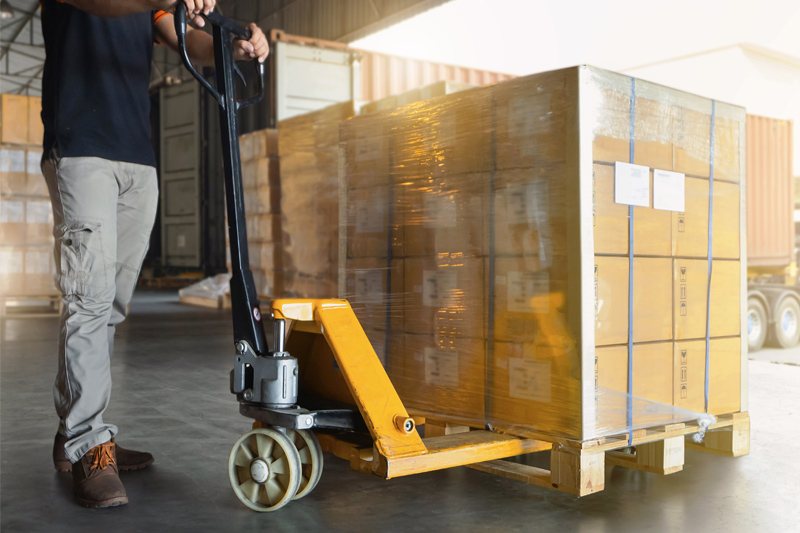 TRABAJADOR EN ALMACÉN DE LOGÍSTICA CON TRANSPALETA CARGADA DE CAJAS.